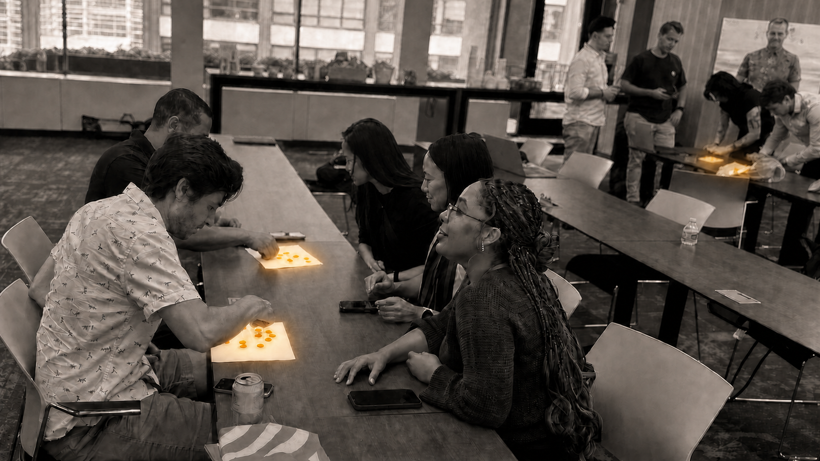 Workshop participants in warm monochrome collaborating at three tables with brightly glowing copper pennies